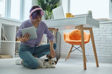 Beautiful young woman in headphones playing with dog in her home office
