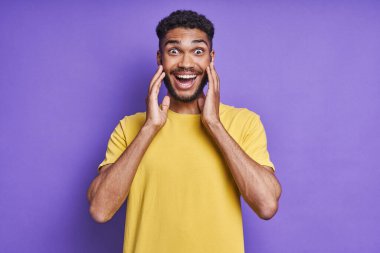 Şaşırmış Afrikalı adam yüzüne yakın el ele tutuşup gülümsüyor ve mor arka planda duruyor.