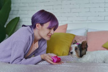 Çekici genç bir kadın köpeğiyle yatakta yatarken elinde küçük bir hediye kutusu tutuyor.