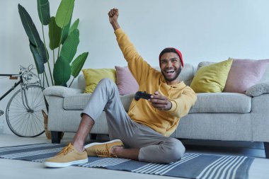 Excited young African man playing video games and gesturing while sitting on the floor at home