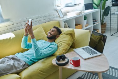Cheerful African man using smart phone while relaxing on the couch at home