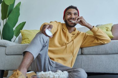 Happy young African man eating popcorn and watching TV while sitting on the floor at home