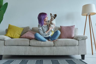Neşeli genç kız evdeki kanepede dinlenirken küçük köpeğini taşıyor.