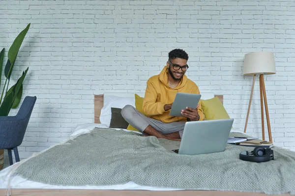 坐在床上的快乐的非洲男人在使用技术 — 图库照片