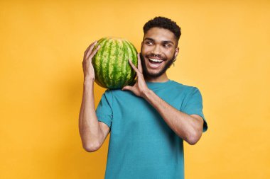 Sarı arka planda dururken omzunda karpuz taşıyan oyuncu Afrikalı adam.