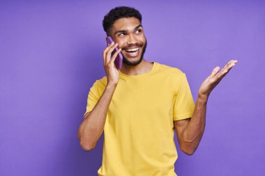 Neşeli Afrikalı adam, mor arka planda dururken cep telefonuyla konuşuyor ve el kol hareketi yapıyor.