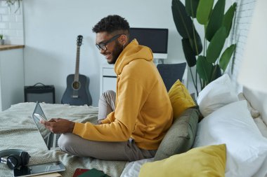 Neşeli Afrikalı adam evde yatakta otururken akıllı telefon kullanıyor.