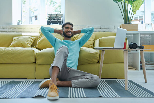 Relaxed African man holding hands behind head and smiling while sitting on the floor at home