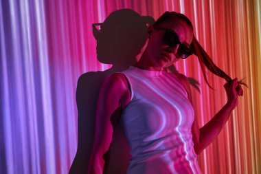 Gözlüklü çekici genç bir kadın. Renkli ışıklarla duvara yaslanırken saçlarını düzeltiyor.