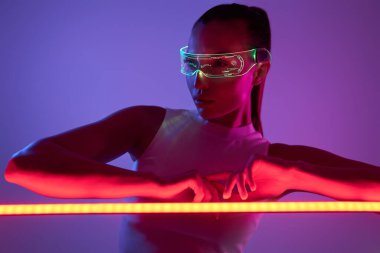 Fütürist gözlüklü çekici bir kadın. Led lambasına yaslanmış karanlık arka plana yaslanıyor.