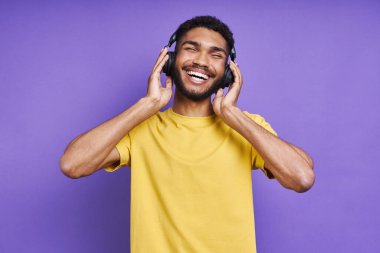 Joyful African man in adjusting his headphones while standing against purple background