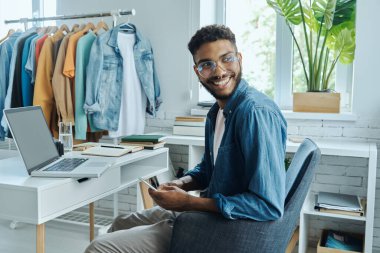 Confident African man holding smart phone and smiling while sitting in fashion store office