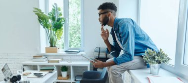 Thoughtful young man holding digital tablet while sitting on the window sill in office