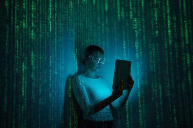 Confident woman in futuristic glasses using digital tablet against dark background with green symbols