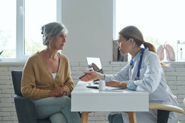 Kendine güveni tam bir kadın doktor. Hastaya bir şey anlatıyor. Tıp bürosunda masada otururken.