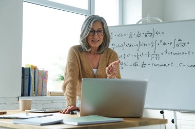 Olgun bir kadın beyaz tahtanın yanında durup dizüstü bilgisayara bakarken matematik öğretiyor.