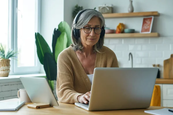 Confident Senior Woman Headphones Using Technologies While Sitting Kitchen Island — Stock Photo, Image