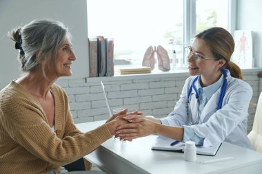 Sağlık ofisinde oturmuş el ele tutuşan kıdemli kadın ve kadın doktor.