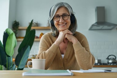 Senior woman in headphones having web conference while sitting at the domestic kitchen