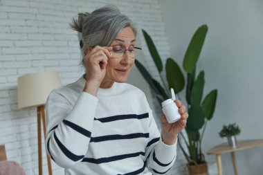 Yaşlı kadın evde otururken elinde ilaçlarla şişenin içine bakıyor.