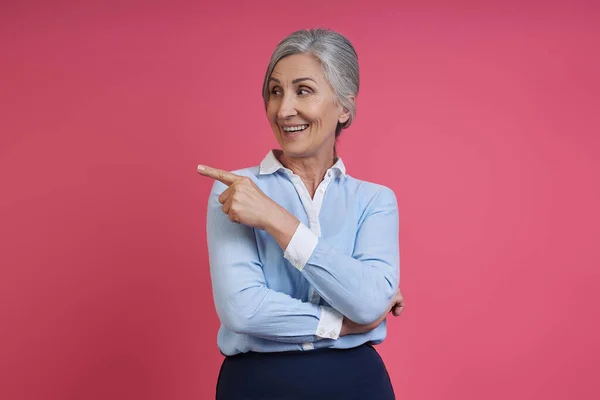 Confident senior woman pointing away and smiling while standing against pink background