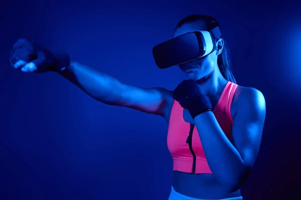 Young woman in virtual reality glasses practicing in punching against colorful background