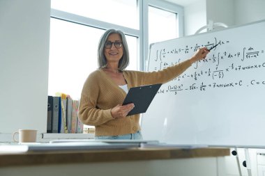 Sınıfta beyaz tahtayı göstererek matematik öğreten neşeli, son sınıf öğrencisi kadın.