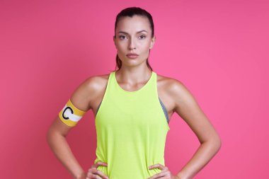 Spor kıyafetleri içinde, kaptan bandı takan ve pembe arka planda duran kendine güvenen bir kadın.