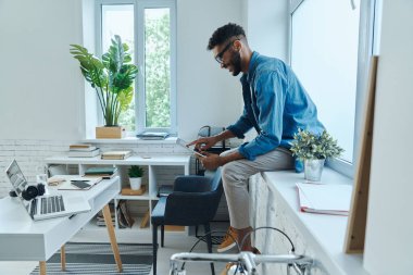 Ofisteki pencere eşiğinde otururken dijital tablet kullanan neşeli Afrikalı adam.