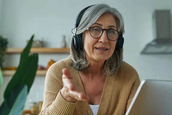 Confident Senior Woman Headphones Gesturing While Sitting Domestic Kitchen — Stock Photo, Image