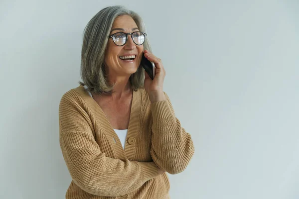 Elegant senior woman talking on mobile phone and smiling while standing against white wall