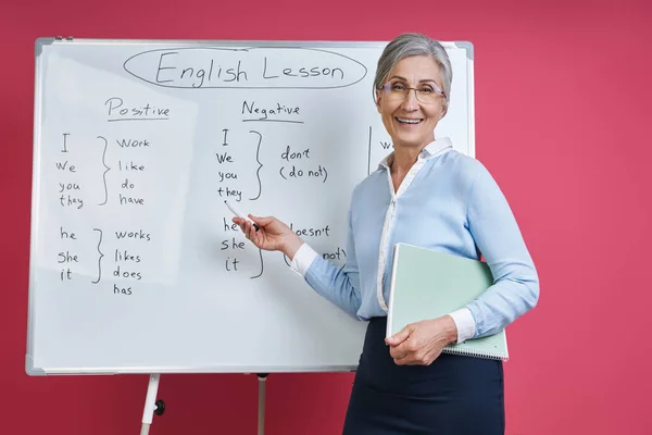 Cheerful senior woman teaching English language near the whiteboard against pink background