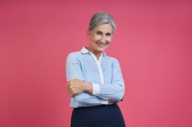 Confident senior woman keeping arms crossed while standing against pink background