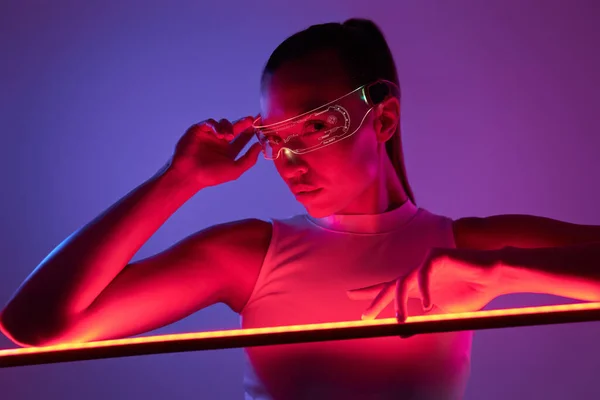 Beautiful woman in futuristic glasses leaning at the LED lamp against dark background