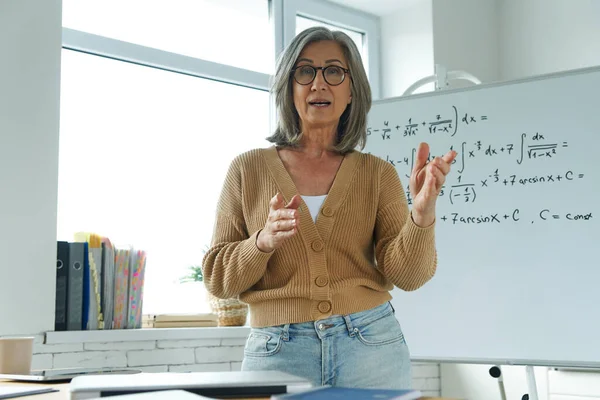 Üst düzey kadın beyaz tahtanın yanında dikilip el kol hareketi yaparken matematik öğretiyor.