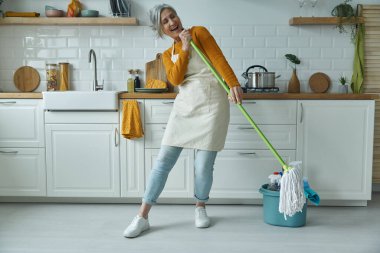 Playful senior woman carrying mop like microphone while standing at the domestic kitchen