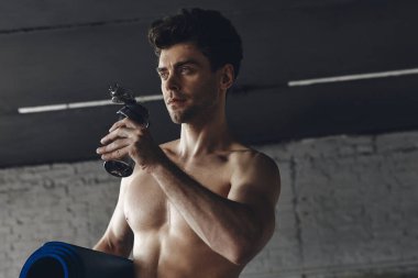 Handsome young man carrying exercise mat and drinking water while standing in gym