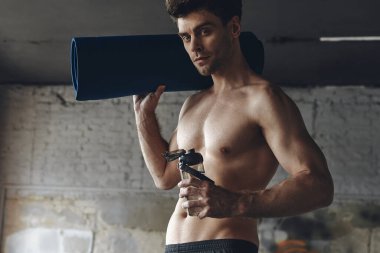 Handsome young man carrying exercise mat and bottle with water while standing in gym