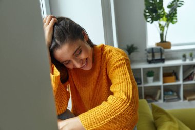 Attractive young woman leaning head on hand and smiling while sitting on the window sill
