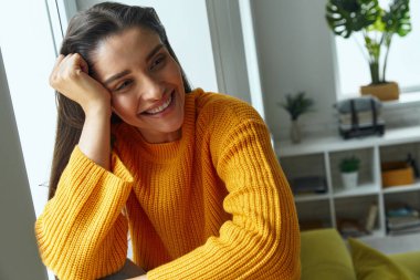 Beautiful young woman leaning head on hand and smiling while sitting on the window sill