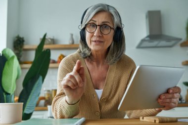 Senior woman in headphones explaining something and gesturing while sitting at the domestic kitchen