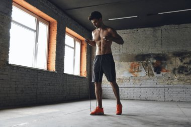 Full length of confident young man stretching jumping rope while standing in gym