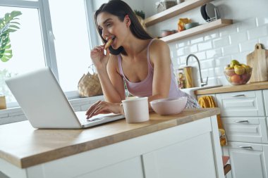 Relaxed young woman using laptop and eating cookies while sitting at the kitchen counter