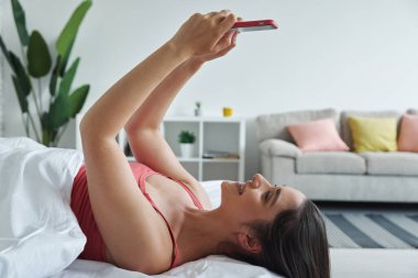 Beautiful young woman using smart phone and smiling while lying in bed at home
