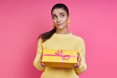 Thoughtful young woman holding gift box and looking away against colored background