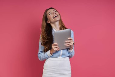 Excited young woman holding digital tablet and keeping eyes closed against colored background