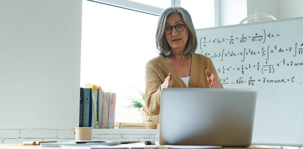 Mature woman teaching mathematics while standing near the whiteboard and looking at laptop