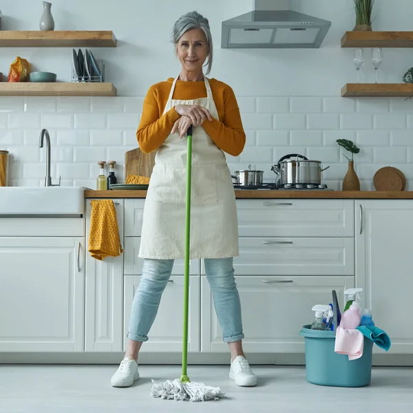 Full length of confident senior woman leaning at the mop while standing at the domestic kitchen