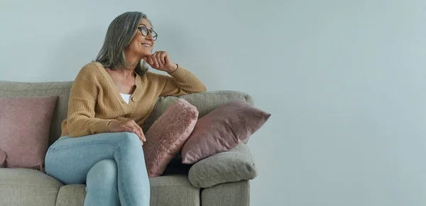 Elegant senior woman holding hand on chin and smiling while relaxing on the comfortable couch