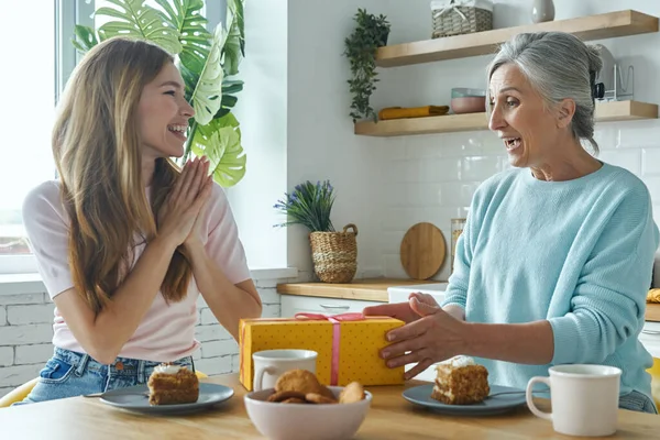 Excited senior mother receiving a gift box from her adult daughter while both sitting at the kitchen